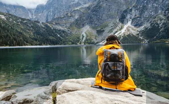 Woman with a yellow raincoat and a black backpack sitting down in the mountains and looks out on a fjord